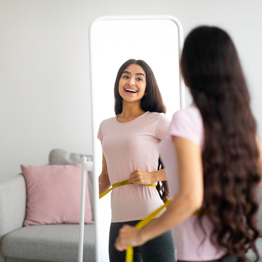 A smiling woman in a pink shirt measures her waist with a tape measure while looking at her reflection in a mirror, with a cozy living room setting in the background.