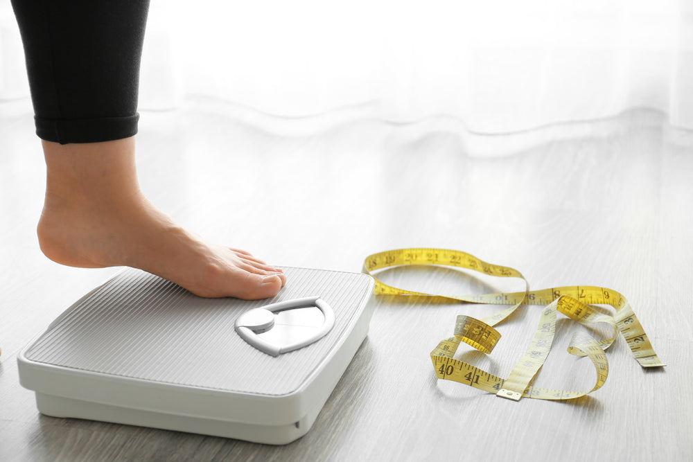 A foot is placed on a bathroom scale, with a measuring tape lying on the floor nearby, suggesting a focus on weight management and progress in a weight loss journey.