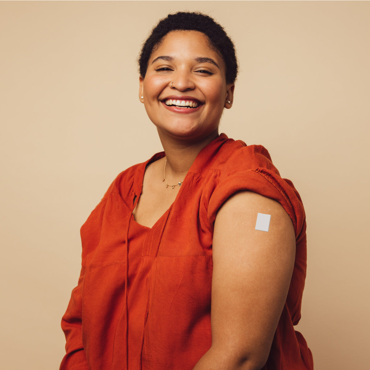 A smiling woman with short hair wears a red shirt and shows her arm with a bandage on it, set against a neutral beige background, suggesting she has recently received a vaccination.
