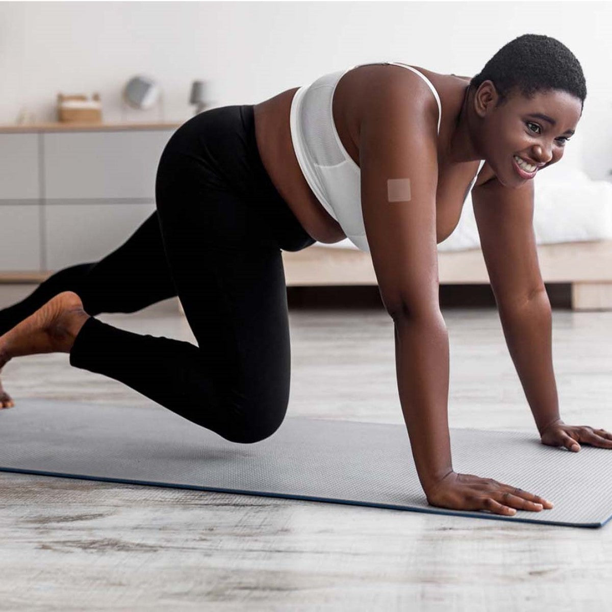 A smiling woman with short hair is exercising on a yoga mat in a bright room, wearing a white sports bra and black leggings, with a vitamin patch visible on her arm.