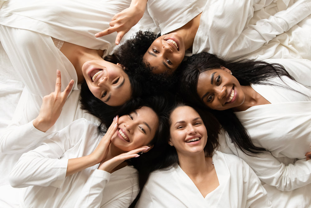 Five women in white robes lie on a bed, smiling and posing playfully, with one making a peace sign, conveying a sense of joy and friendship in a cozy, relaxed setting.