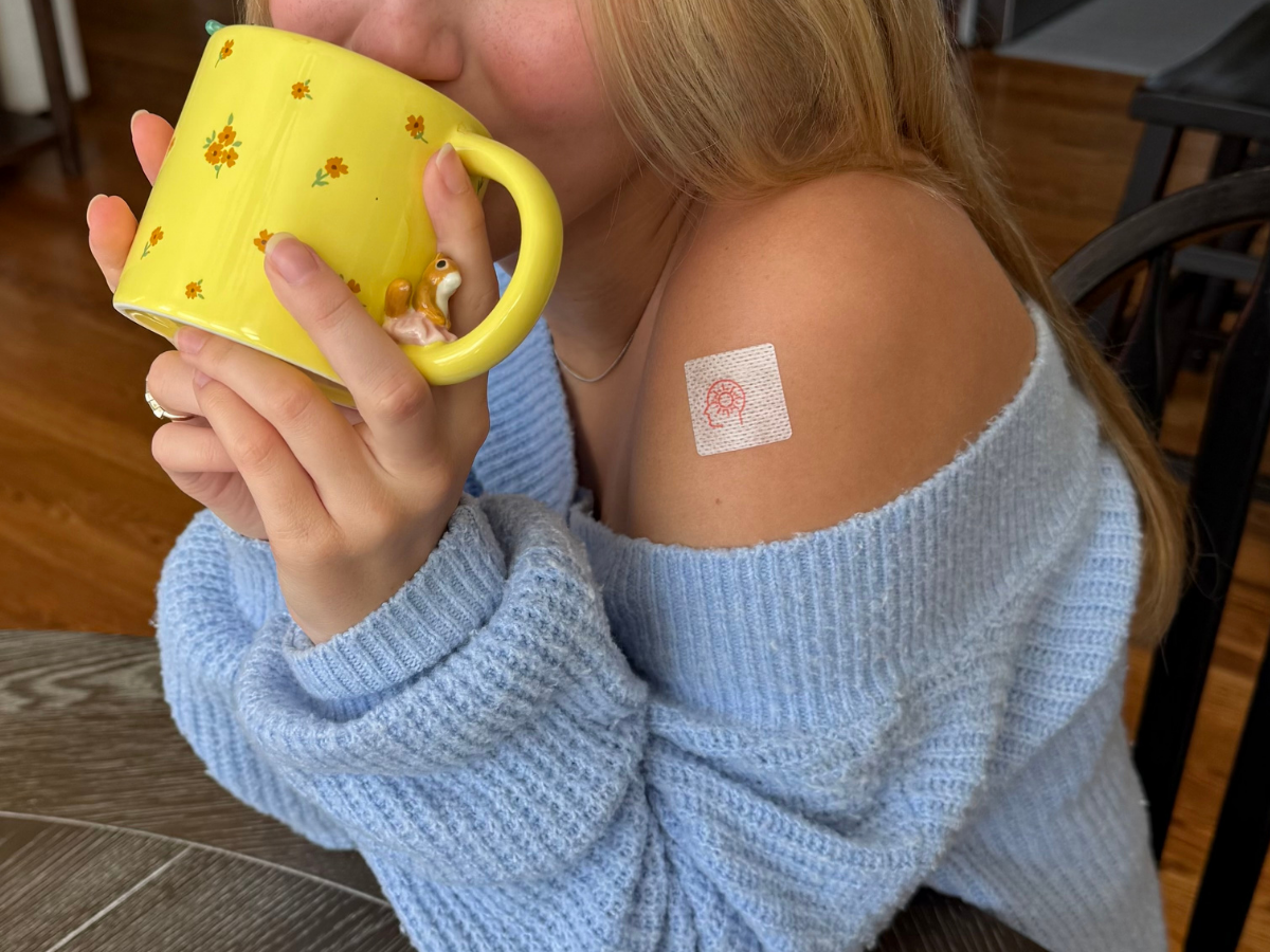 A person wearing a light blue off-the-shoulder sweater drinks from a yellow mug with a squirrel figure, displaying a square medical patch with a red design on their shoulder.