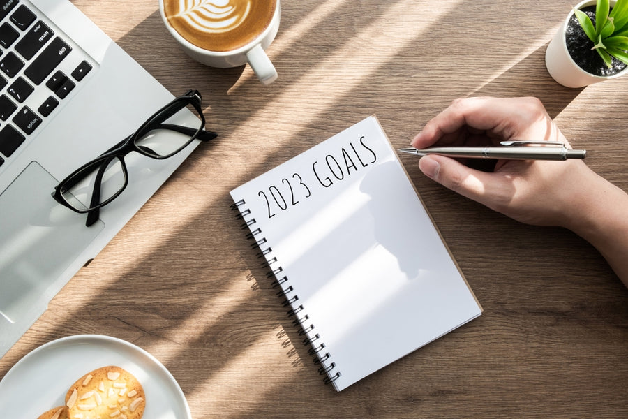 A hand holds a pen poised over a notebook labeled "2023 GOALS," with a laptop, glasses, a cup of coffee, and a plate of cookies on a wooden table, suggesting a planning or goal-setting session.