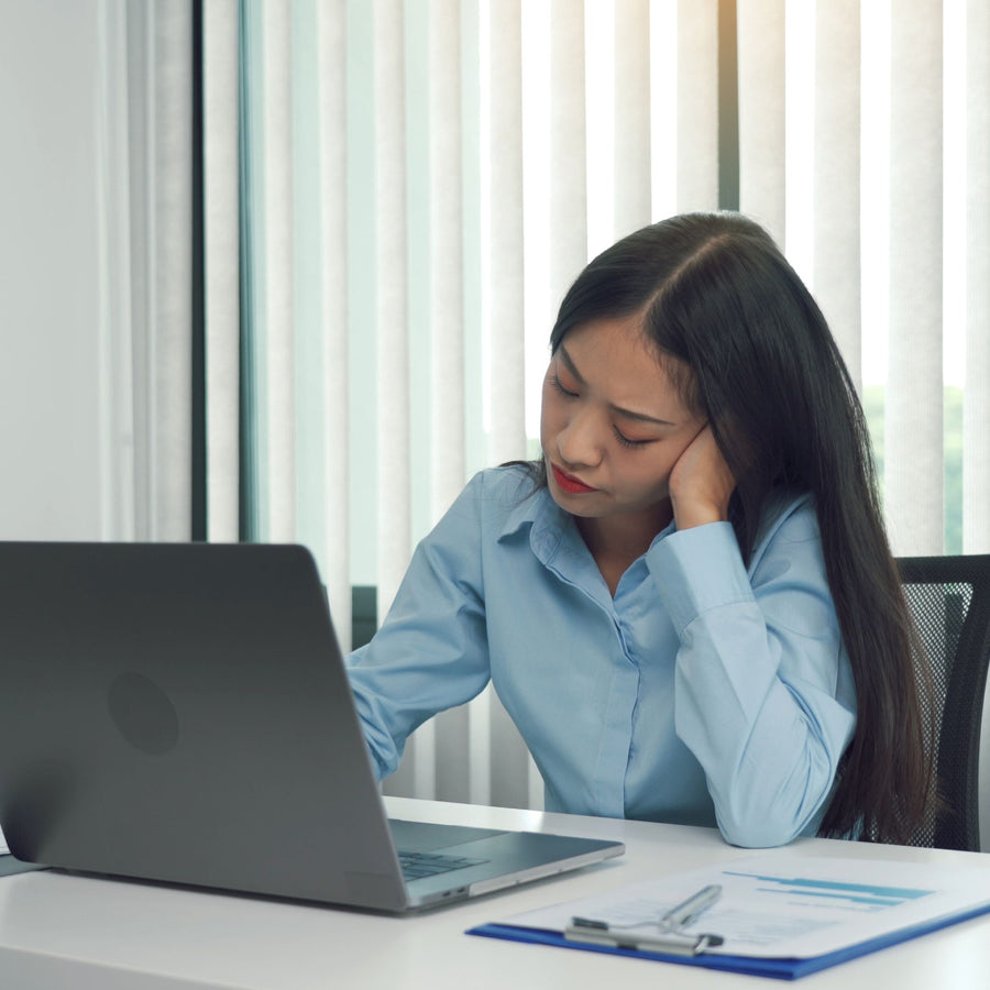 A woman in a blue shirt rests her head on her hand while looking at a laptop, appearing tired and distracted in a bright office with vertical blinds and a clipboard nearby.