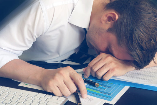 A man in a white shirt rests his head on a cluttered desk covered with papers and a keyboard, showing signs of exhaustion, possibly related to stress or fatigue.
