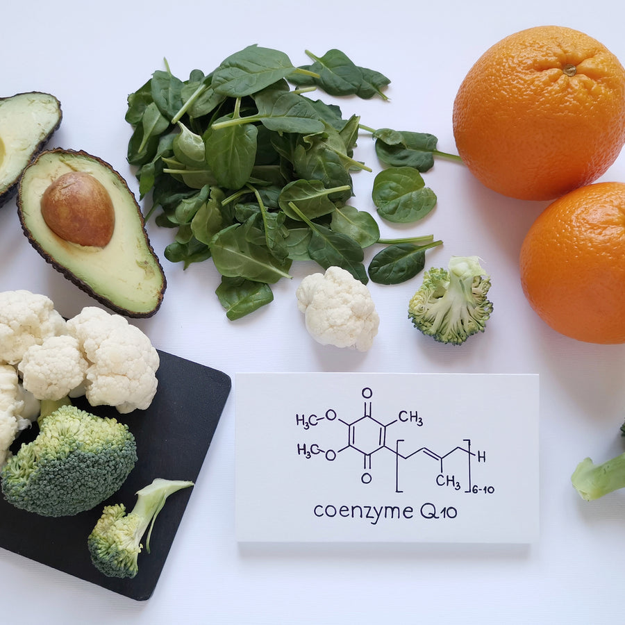 A variety of fresh vegetables, including avocado, spinach, cauliflower, and broccoli, are arranged alongside two oranges and a card displaying the chemical structure of coenzyme Q10.