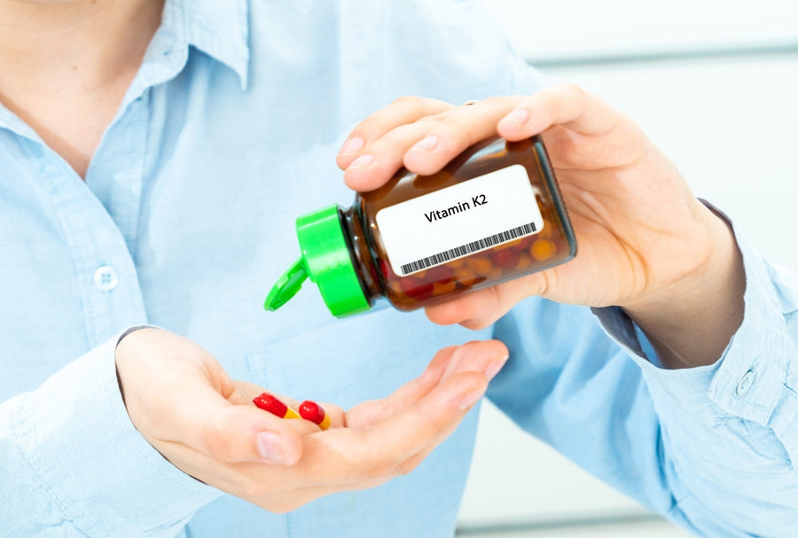 A person in a light blue shirt is holding a bottle labeled "Vitamin K2" while pouring red and yellow capsules into their other hand, suggesting a focus on health and supplementation.