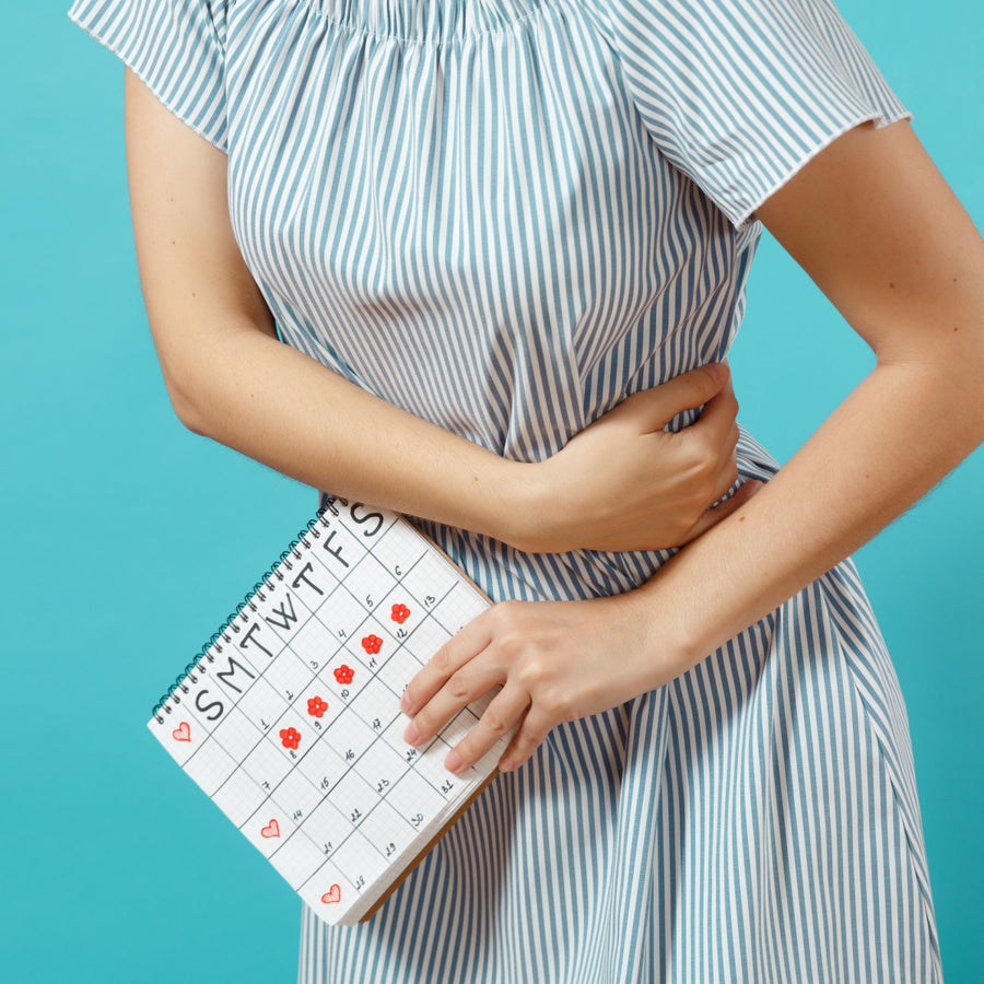 A person in a striped dress holds a calendar marked with flowers and hearts while gently cradling their abdomen, suggesting discomfort related to PMS symptoms against a bright blue background.