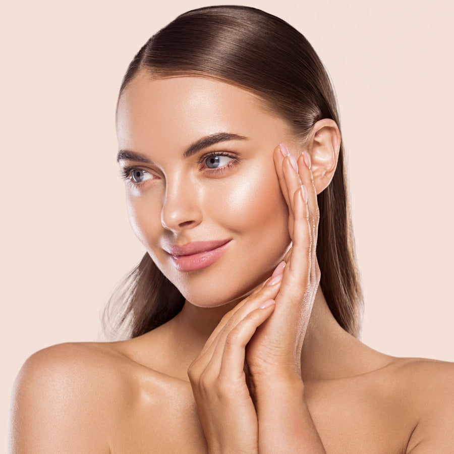A woman with smooth skin and a gentle smile touches her face with one hand, set against a soft, neutral background that highlights her natural beauty.