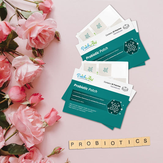 Three PatchAid Probiotic Patch packages are arranged on a light pink surface with pink roses nearby, and wooden letters spell out "PROBIOTICS" beneath them, highlighting gut health.