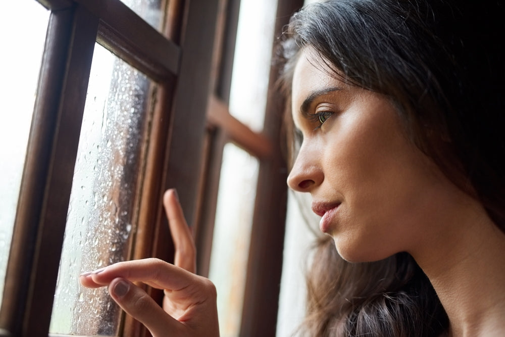 A woman with long dark hair gazes thoughtfully out of a rain-streaked window, resting her fingers on the glass, reflecting a mood of contemplation during a gloomy day.