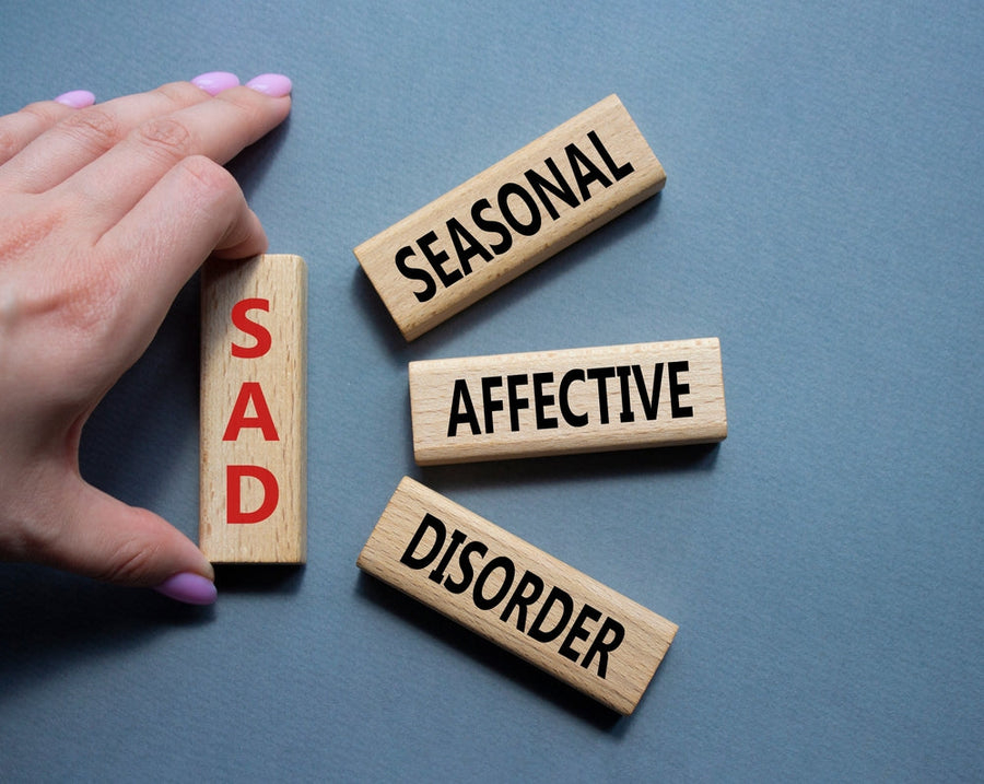 A hand holds a wooden block labeled "SAD" in red, while three other blocks reading "SEASONAL," "AFFECTIVE," and "DISORDER" are arranged nearby on a blue background, illustrating the concept of Seasonal Affective Disorder.