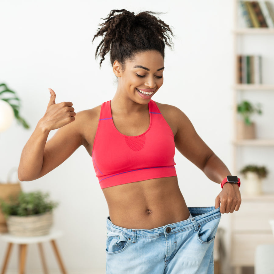 A smiling woman in a pink sports bra gives a thumbs-up while holding up oversized jeans, standing in a bright, modern room with plants and shelves in the background, celebrating her weight loss success.