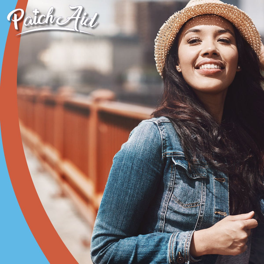 A smiling woman in a denim jacket and straw hat stands on a sunny day by a railing, enjoying the outdoors while promoting PatchAid.