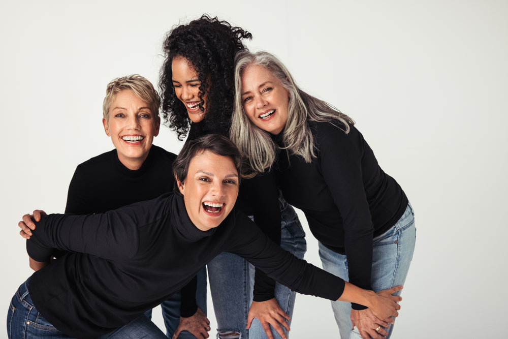 Four women of diverse ages and hairstyles, dressed in black tops and jeans, are joyfully posing together, smiling and laughing in a bright, neutral setting, celebrating friendship and healthy aging.