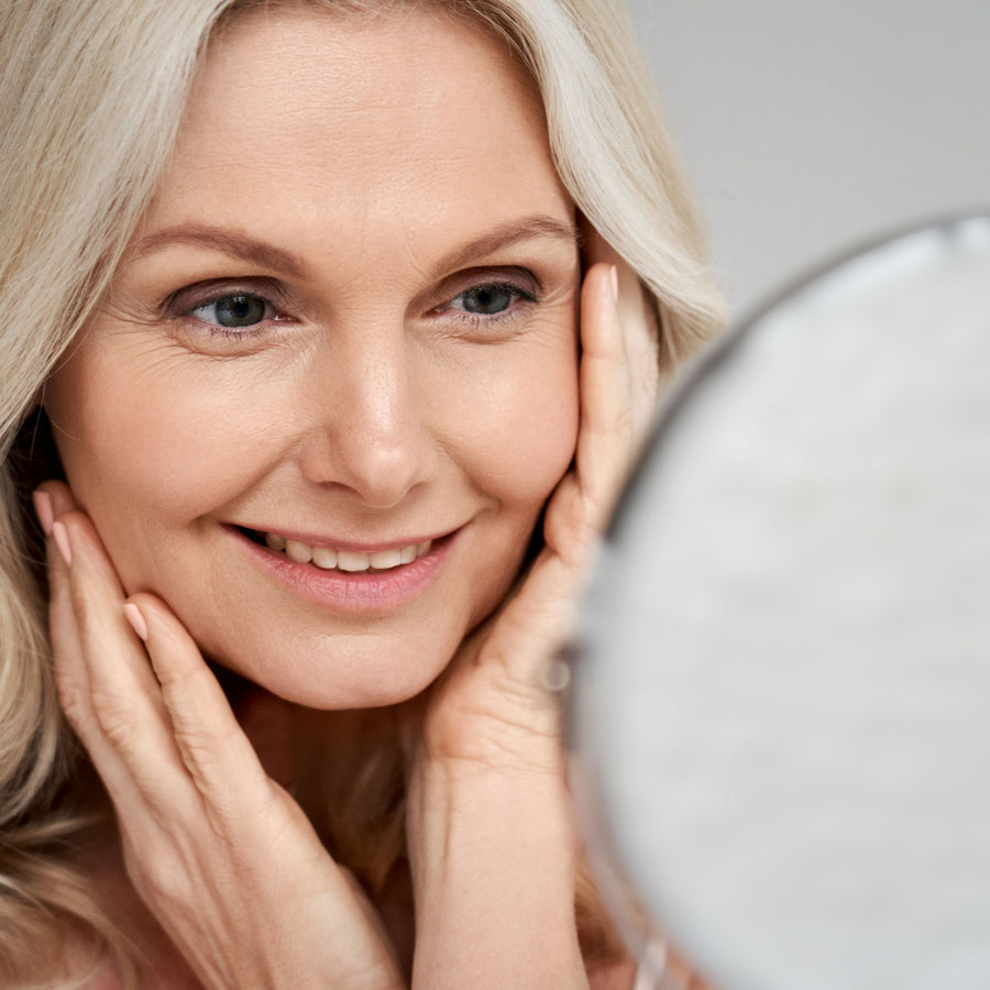 A smiling older woman gently touches her face while looking into a mirror, reflecting a moment of self-care and confidence in a bright, neutral setting.