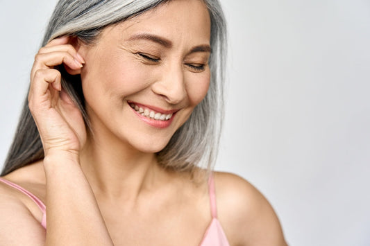 A smiling older woman with long, gray hair gently touches her ear while closing her eyes, conveying a sense of joy and contentment against a soft, neutral background.