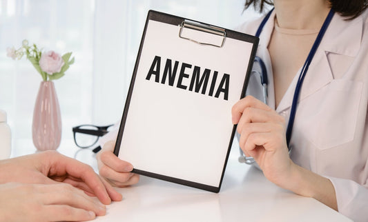 A healthcare professional holds a clipboard displaying the word "ANEMIA" while sitting at a table, with a patient's hands visible, in a well-lit medical office setting.