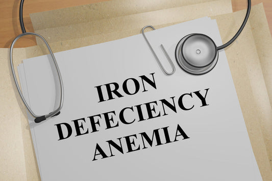 A stethoscope rests on a sheet of paper with the bold title "IRON DEFICIENCY ANEMIA," surrounded by other documents on a wooden surface, suggesting a medical context.
