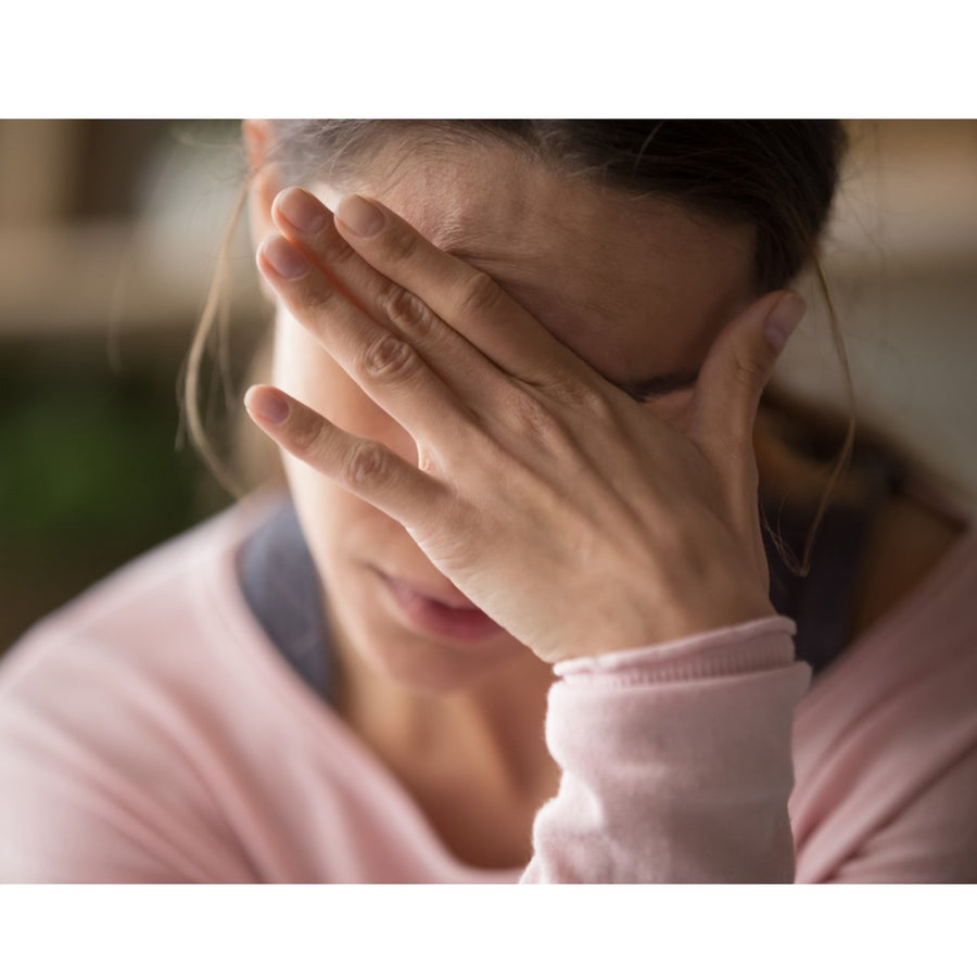 A woman in a pink sweater sits with her head down, covering her face with her hand, conveying a sense of distress or anxiety in a softly lit indoor setting.