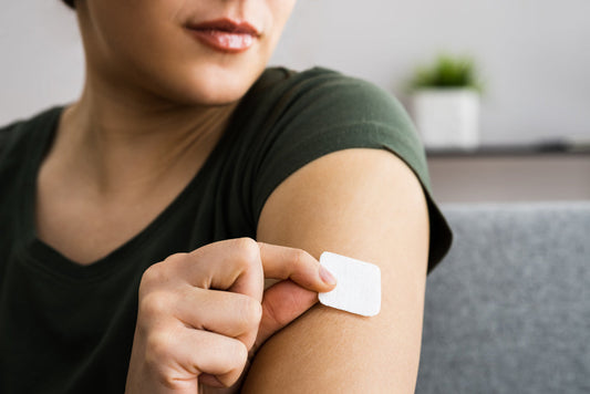 A person in a green shirt applies a vitamin patch to their upper arm while sitting on a couch, highlighting the convenience of using patches over traditional supplements.