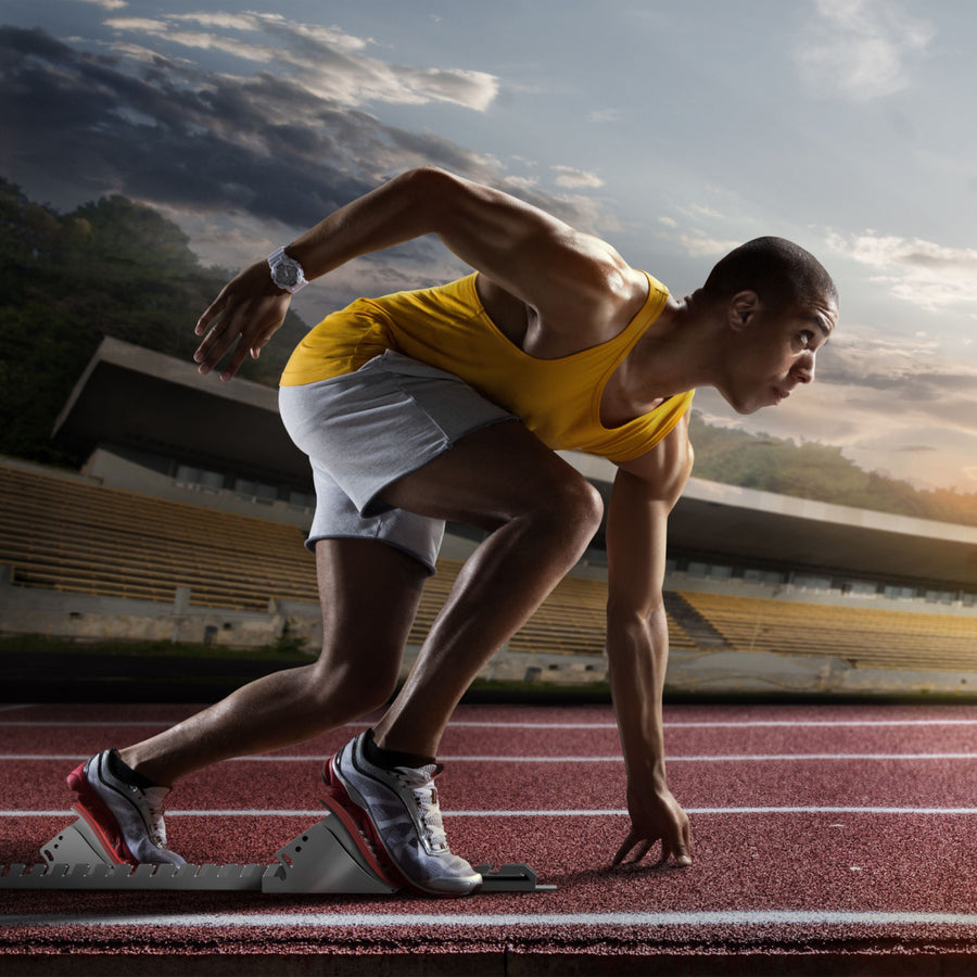 A focused athlete in a yellow tank top prepares to sprint from starting blocks on a track, showcasing determination against a backdrop of a stadium and cloudy sky.
