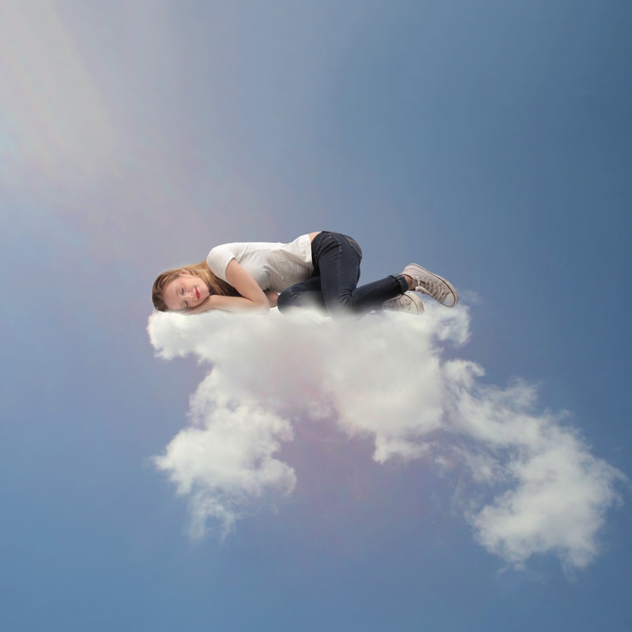 A young woman in casual clothing is curled up and sleeping peacefully on a fluffy cloud against a bright blue sky.