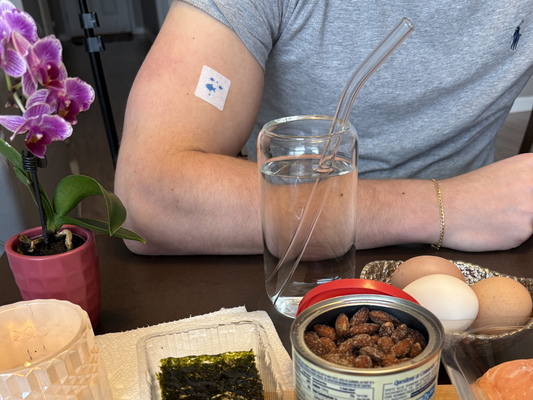 Person wearing a PatchAid vitamin patch on their upper arm while sitting at a table with a glass of water, healthy snacks (eggs, almonds, seaweed), and a glass straw, highlighting a simple daily wellness routine.