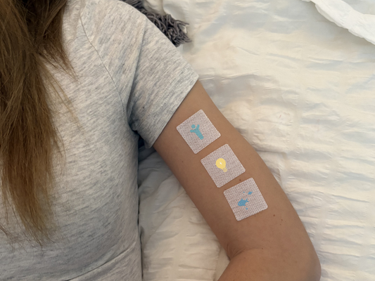Person resting on a bed wearing three vitamin patches on their upper arm, representing daily wellness support.
