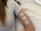 Person resting on a bed wearing three vitamin patches on their upper arm, representing daily wellness support.