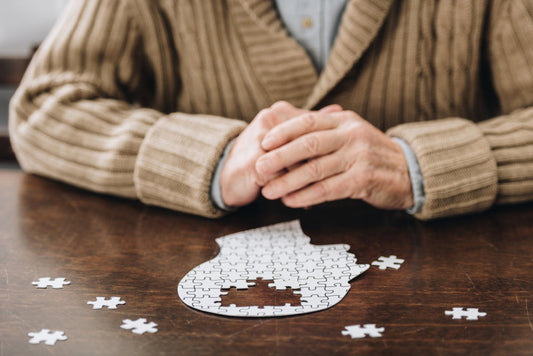 An older adult in a cozy sweater sits at a wooden table, hands clasped, with a partially completed jigsaw puzzle and scattered pieces nearby, suggesting focus on cognitive activities.