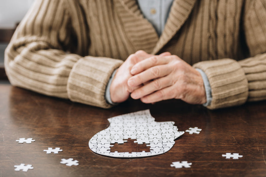 An older adult in a cozy sweater sits at a wooden table, hands clasped, with a partially completed jigsaw puzzle and scattered pieces nearby, suggesting focus on cognitive activities.