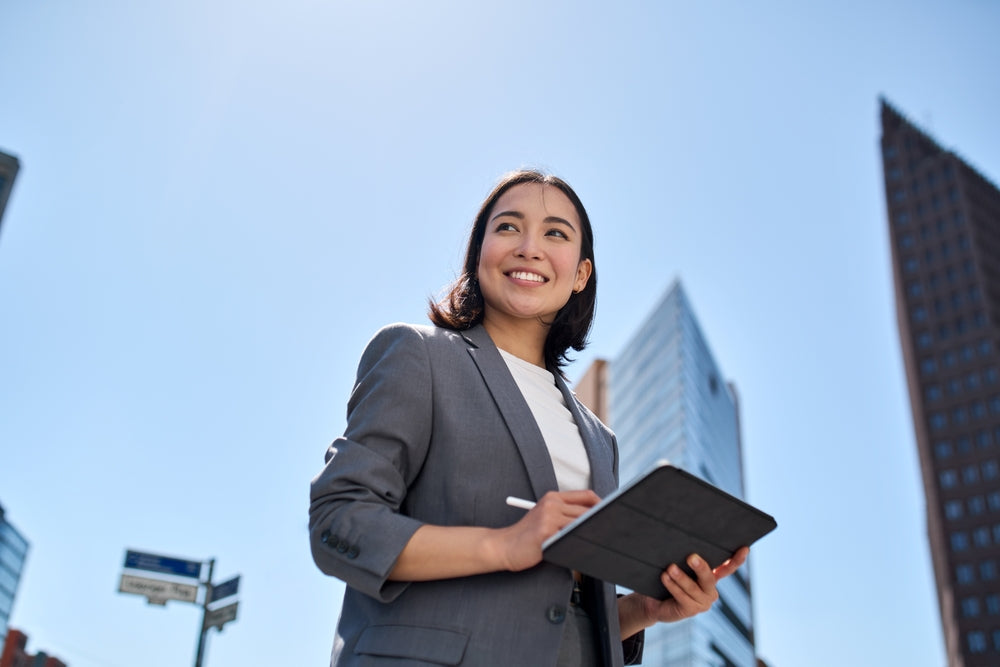 A professional woman in a gray suit smiles while holding a tablet, standing outdoors against a backdrop of tall buildings under a clear blue sky.