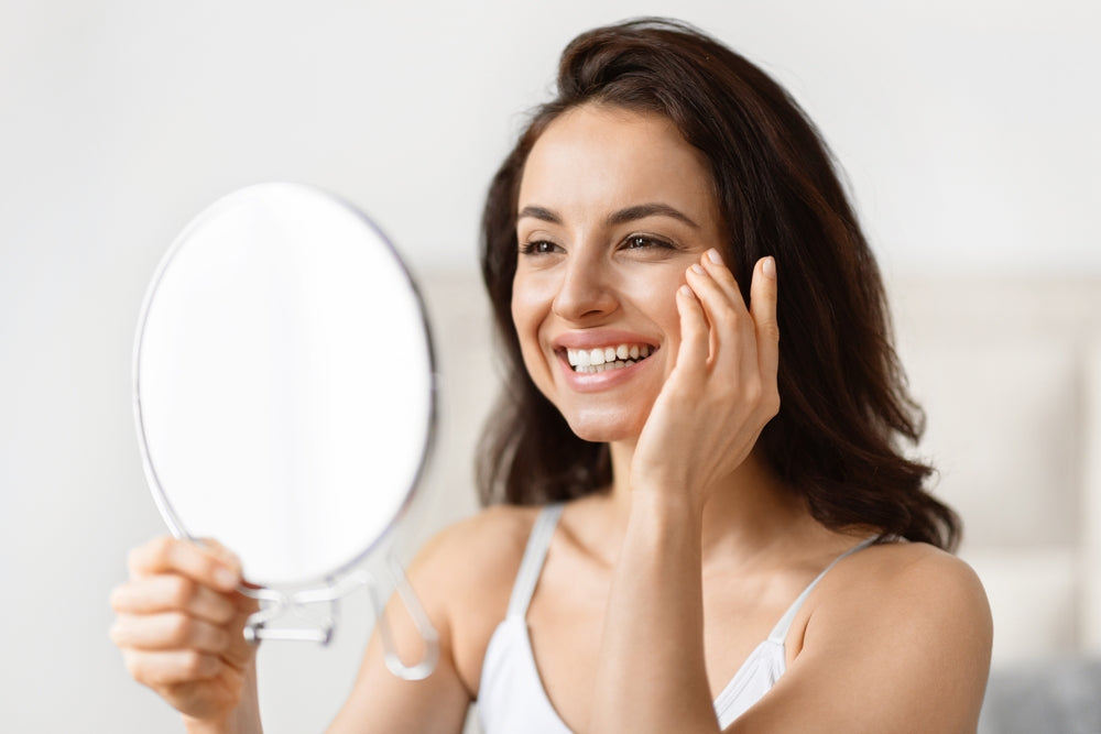 A woman smiles while looking in a mirror and touching her face, suggesting a positive skincare routine, in a bright, softly lit room.