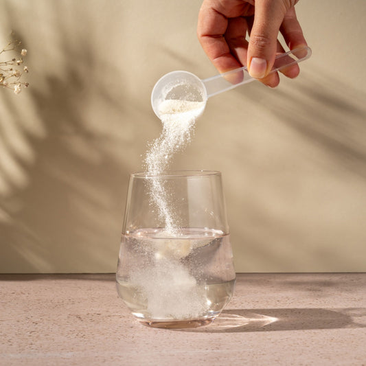 A hand pours collagen powder from a measuring scoop into a glass of water on a light-colored surface, with soft shadows and a neutral background enhancing the scene.
