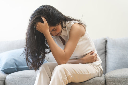 A woman sits on a couch, leaning forward with her head in one hand and her other hand on her abdomen, expressing discomfort related to menstrual pain in a light-filled room.