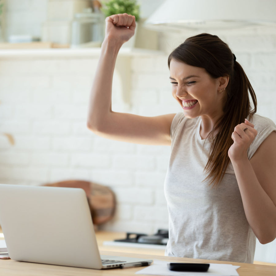 A young woman with a ponytail celebrates excitedly at her desk, raising her fists in joy while looking at her laptop in a bright, modern kitchen.