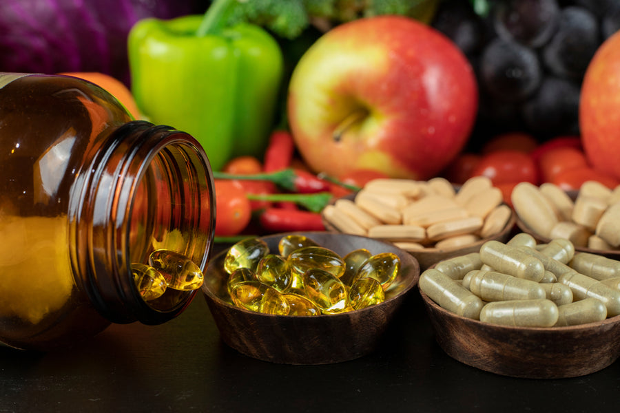 A brown bottle spills yellow gel capsules onto a dark surface, surrounded by bowls of various supplements and a colorful assortment of fresh fruits and vegetables in the background.