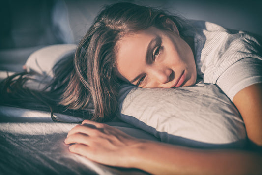 A woman with long hair rests her head on a pillow, looking tired and contemplative, in a softly lit bedroom setting, suggesting feelings of fatigue or low energy.