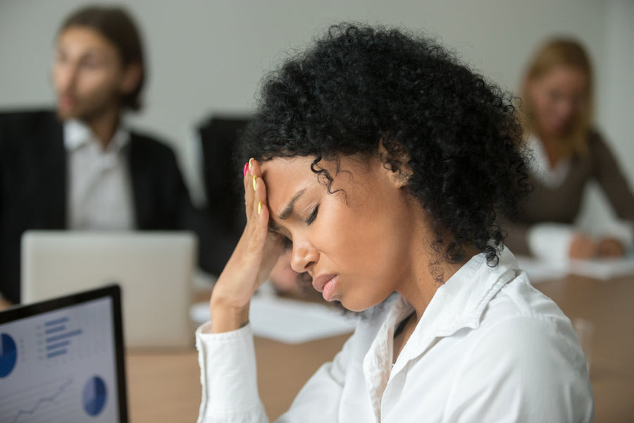 A woman with curly hair looks stressed and holds her forehead while sitting at a conference table, with colleagues in the background working on laptops and papers.