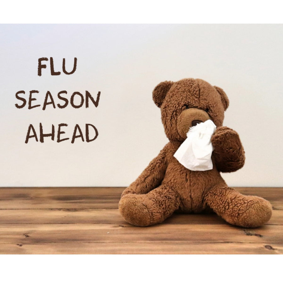 A brown teddy bear sits on a wooden surface, holding a tissue to its nose, with the text "FLU SEASON AHEAD" displayed beside it, emphasizing the need for flu season preparation.