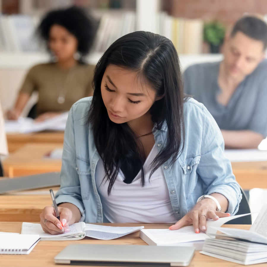 A student with long dark hair focuses intently on writing in a notebook while surrounded by books and classmates in a bright, modern study space.