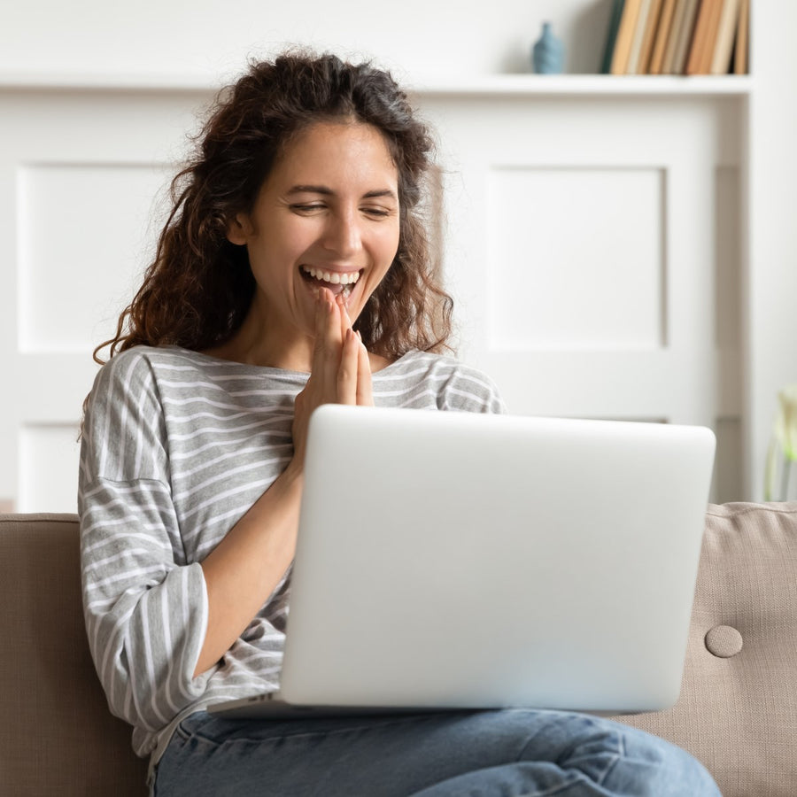 A young woman with curly hair smiles joyfully while sitting on a couch, looking at her laptop with her hands clasped in excitement, in a bright, cozy living room.