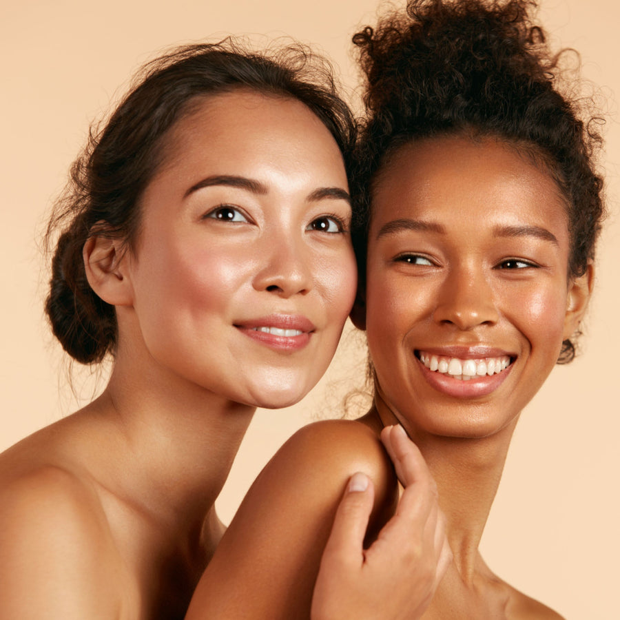 Two women with glowing skin smile at the camera, one with her arm around the other's shoulder, set against a soft, warm beige background.
