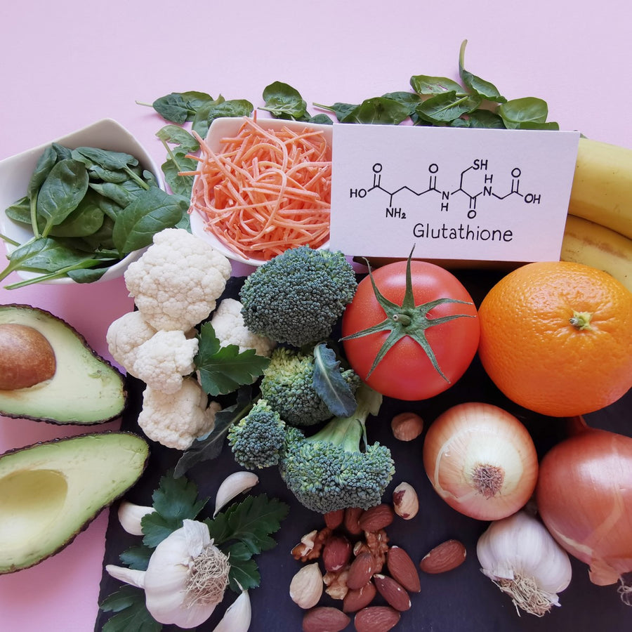 A variety of fresh vegetables and fruits, including broccoli, cauliflower, spinach, tomatoes, and bananas, are arranged with a card displaying the chemical structure of glutathione on a pink background.