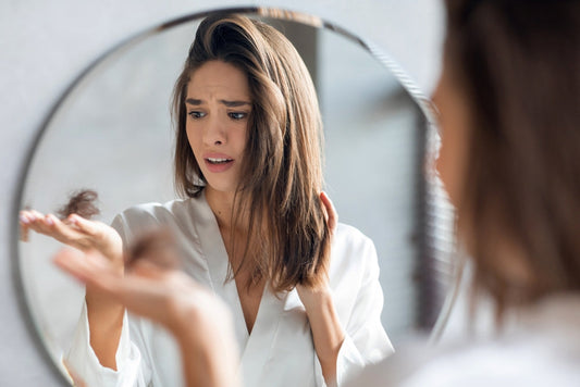 A woman in a white robe looks worried while examining a handful of hair in front of a mirror, reflecting her concern about temporary hair loss in a well-lit bathroom setting.
