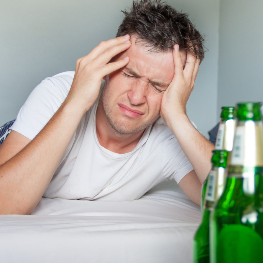 A young man in a white shirt sits on a bed, holding his head with a pained expression, while several empty green beer bottles are visible nearby in a simple room.