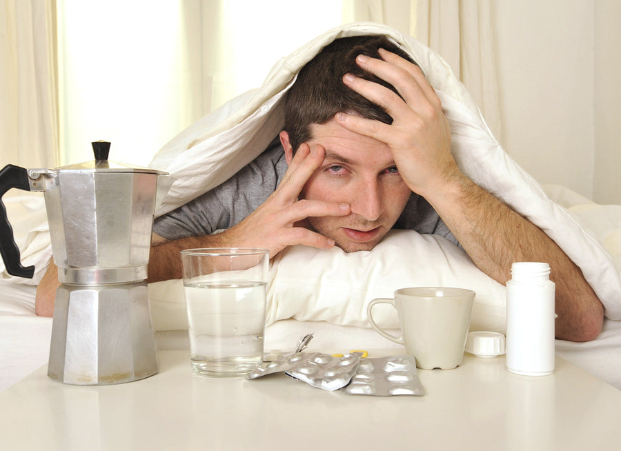 A man sits in bed with a pained expression, holding his head, surrounded by a coffee maker, glasses of water, a cup, and medication, indicating he is dealing with a hangover.