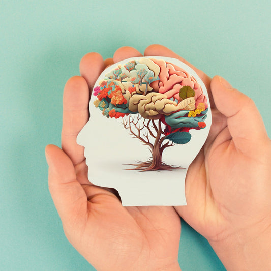 A pair of hands hold a cut-out silhouette of a head, featuring a colorful brain intertwined with flowers and leaves, set against a light blue background, symbolizing mental health and nutrition.