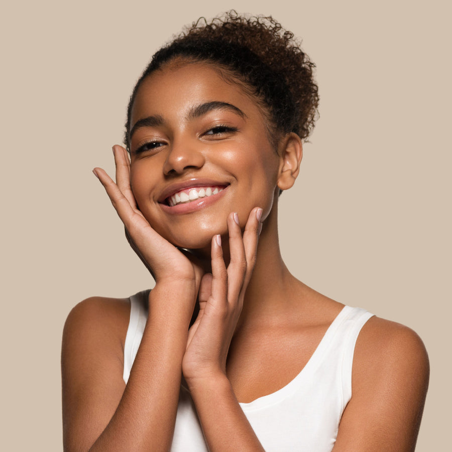 A young woman with curly hair smiles brightly while gently touching her face, set against a soft beige background, highlighting her radiant skin.